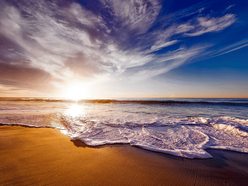 Waves reaching a sandy beach with the sun low near the horizon and clouds across the sky.