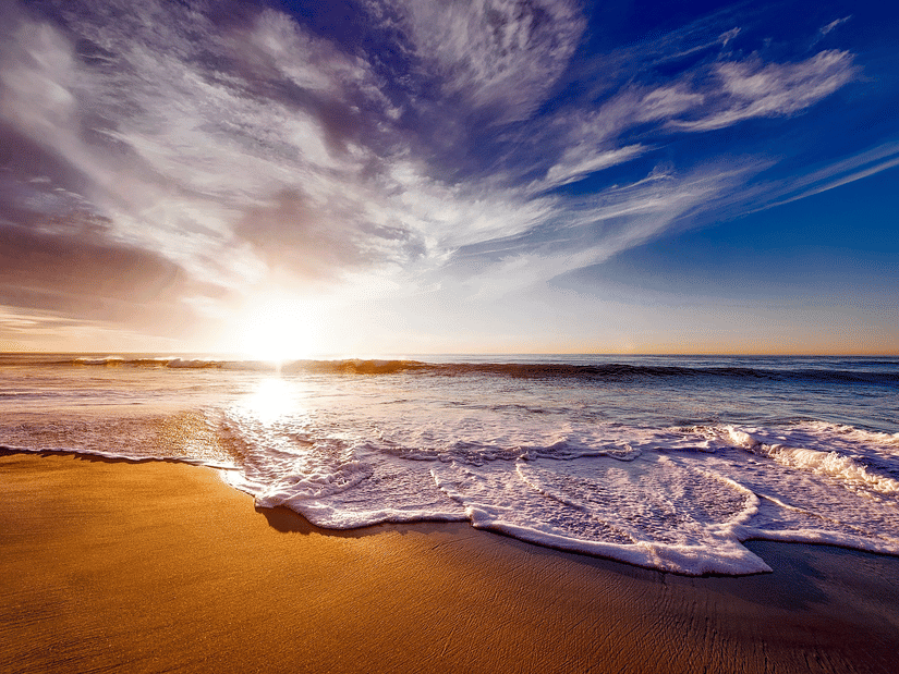 Waves reaching a sandy beach with the sun low near the horizon and clouds across the sky.