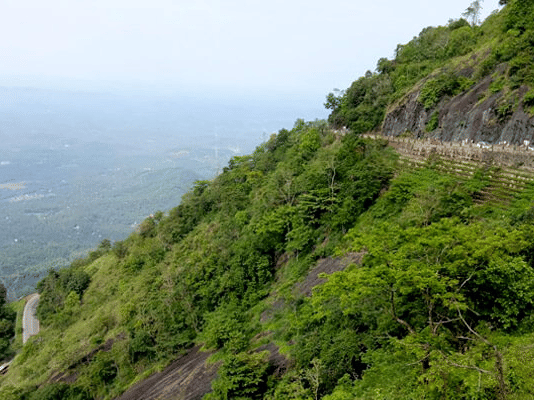 A winding mountain road carved into a steep, lush green cliff side overlooking a vast, misty valley.