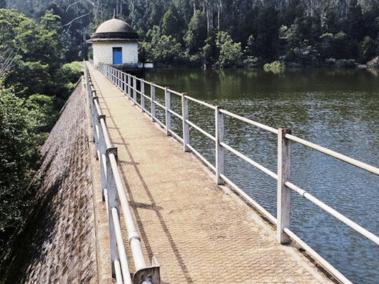 A dam with a walkway along the side, surrounded by dense forested hills and water below, signifying Aliyar Dam, one of the best places to see in Coimbatore.