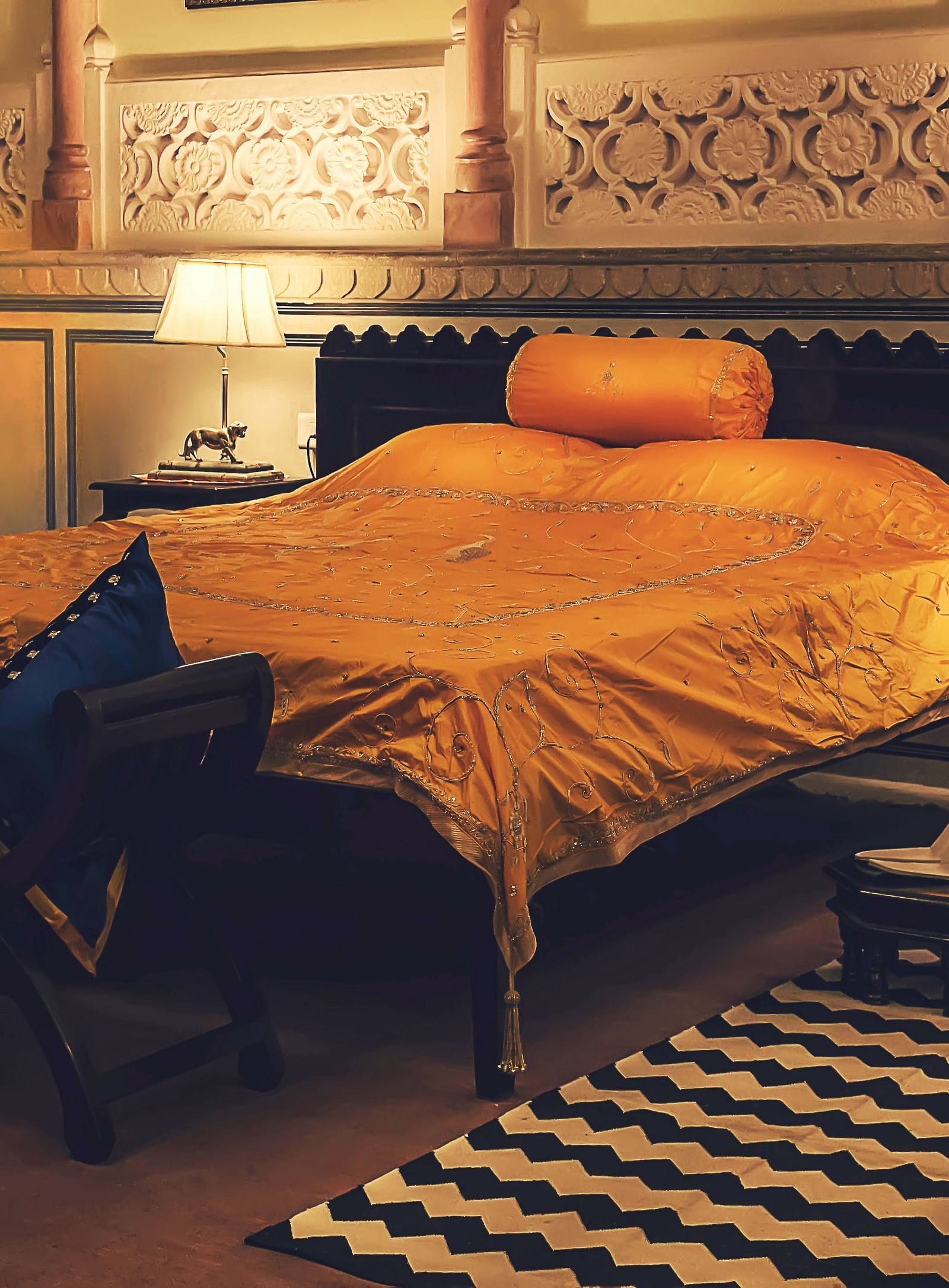 A bedroom at Umaid Vilas Jaipur with a bed dressed in golden fabric, two lamps, and decorative wall cutouts.