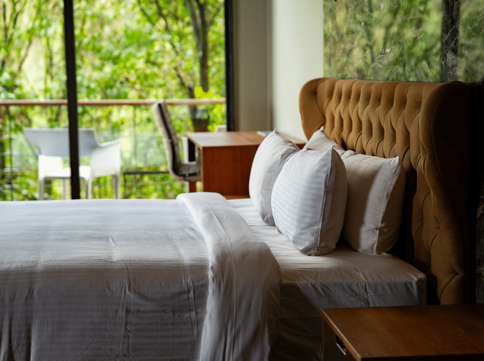 A close-up of a large bed with a high, tufted headboard at Stanley Revelation, with a forest visible outside the window.