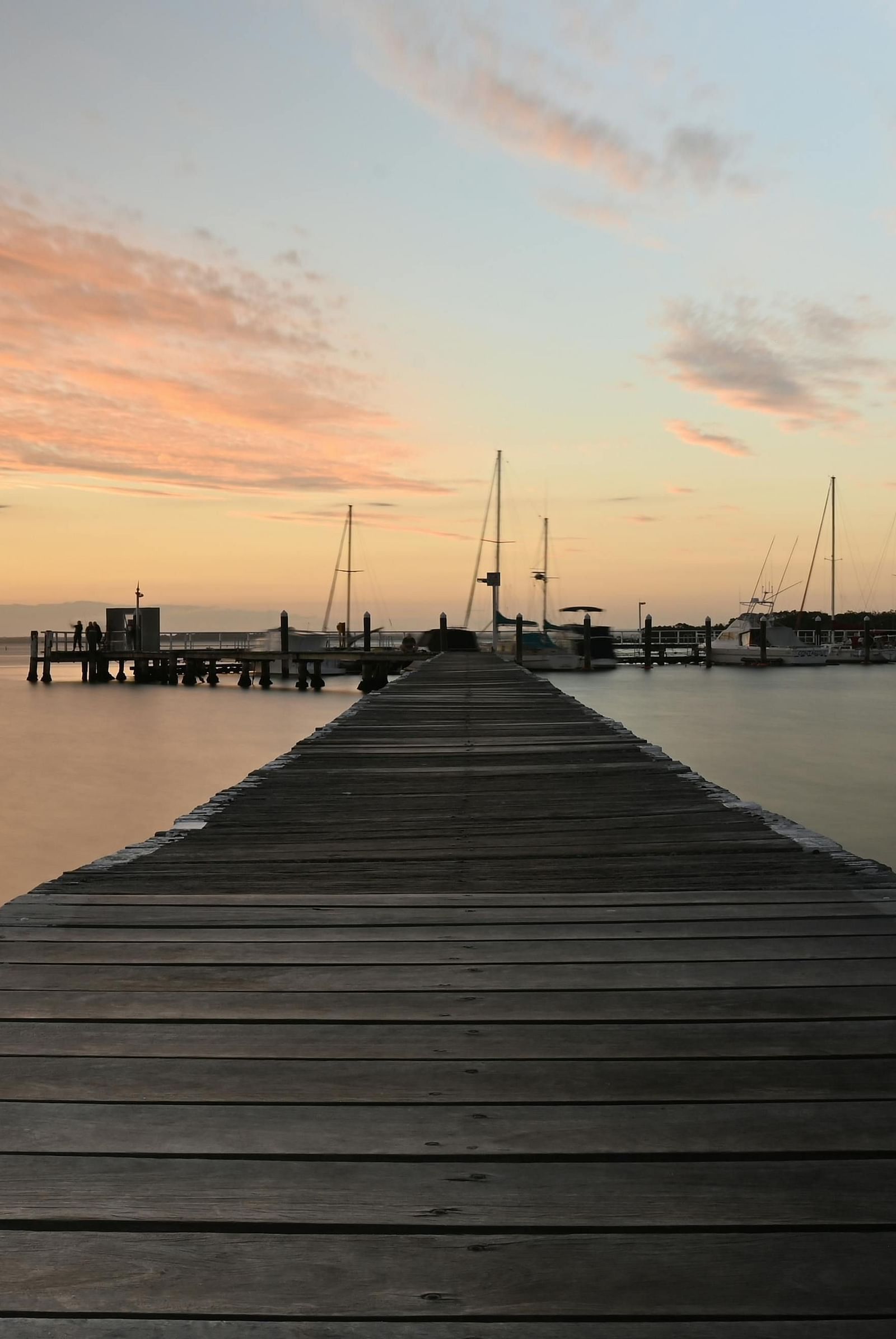 A wooden pier extending into a calm bay, with a beautiful sunset and moored sailboats in the background