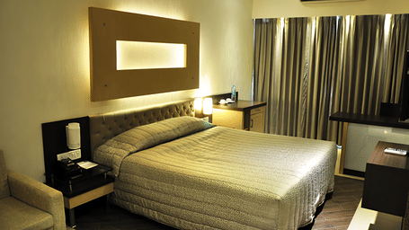 View of a room at Hotel Mumbai House with a king bed, backlit headboard panel, dark curtains, and warm golden ambient lighting.