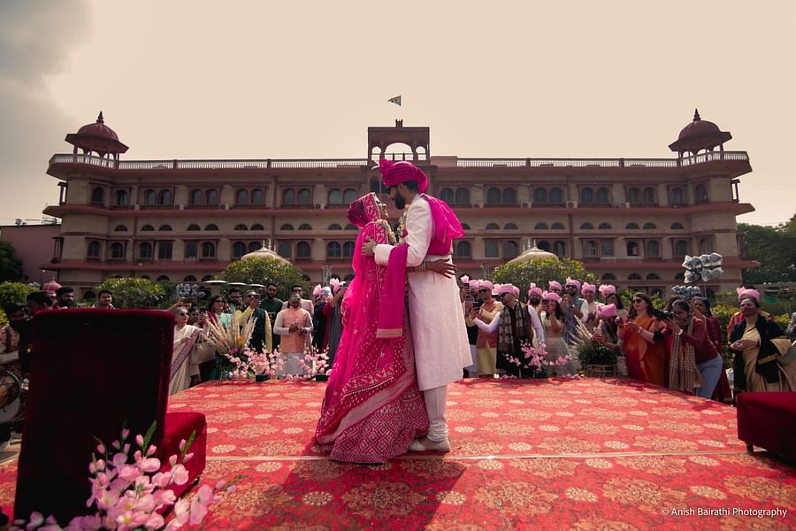 A newlywed couple in wedding attire shares an intimate embrace on a decorated stage, surrounded by cheering guests at a Destination Wedding Resort in Jaipur - Umaid Palace - A Lakeside Heritage Palace Resort
