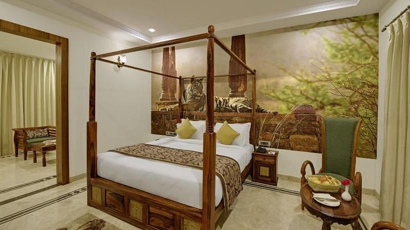 A luxurious four-poster bed takes center stage in this warmly lit room, enhanced by a captivating tiger mural and elegant furnishings, suggesting a stay at Clarks Safari Ranthambore