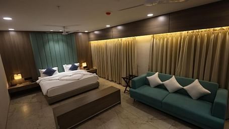 A cosy suite with a king size bed, blue sofa with cushions, bedside tables with lamps, and curtains expanding across the back of the sofa to the bed at Lords Eco Inn Navsari.