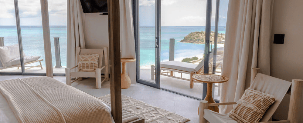 A bright, airy bedroom with a large bed and a view of the ocean through an open doorway, suggesting a beachfront resort.