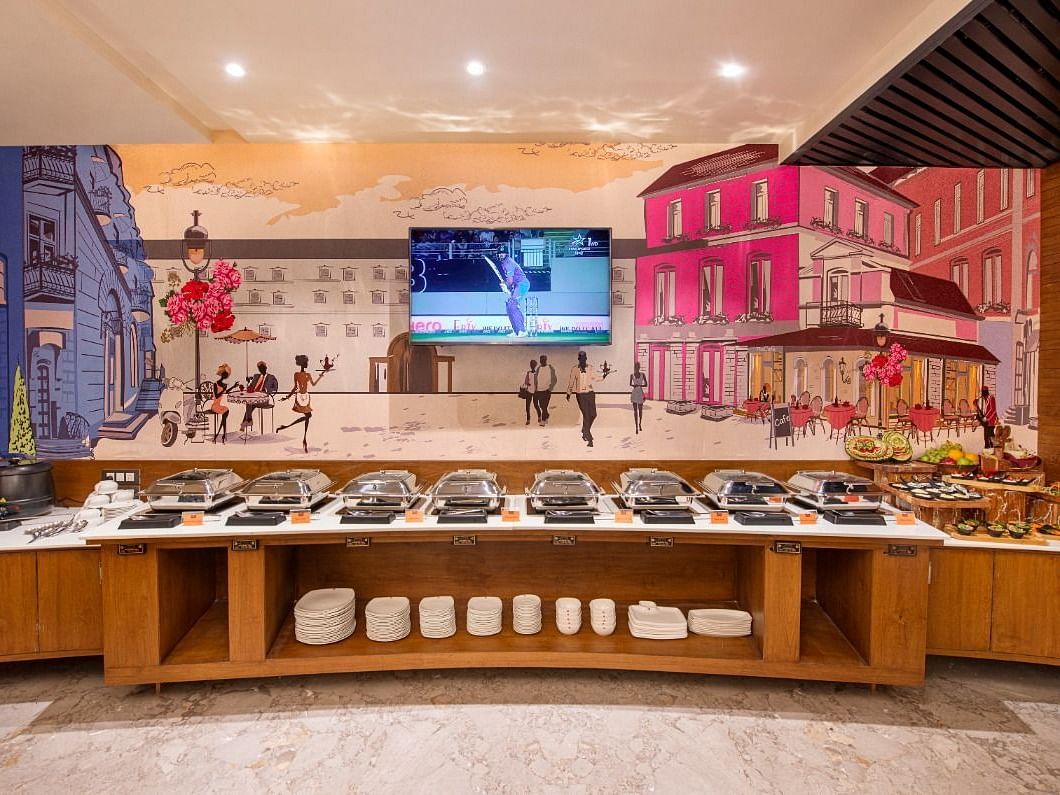 A buffet spread is presented on a long wooden counter with various covered dishes and white containers, set against a backdrop of a colourful mural depicting a European street scene and a television screen at Lords Inn Motikhavdi, Jamnagar.