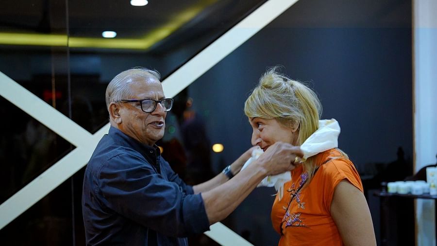 A man with glasses adjusts a white garment around a woman's neck in a salon or similar setting.