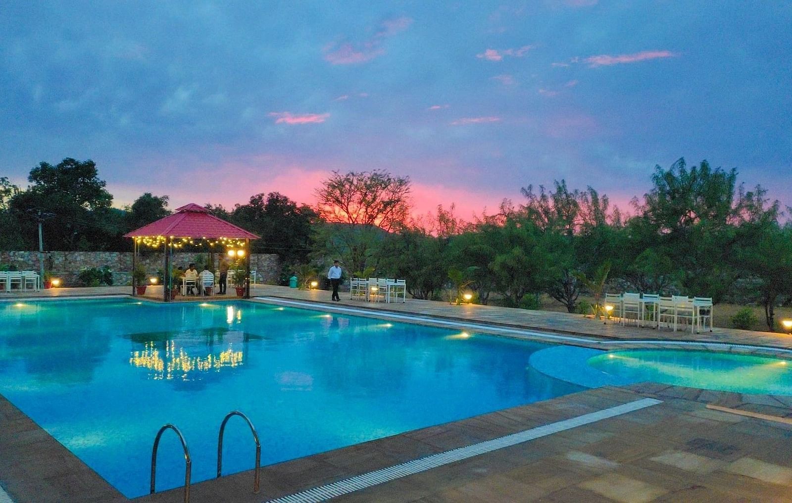 An outdoor swimming pool at dusk with a pink and blue sky in the background - Bamboo Saa Mulberry Resort, Pushkar