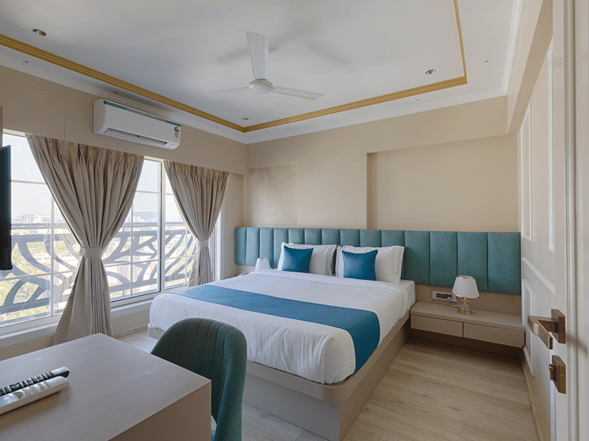 A well-lit deluxe bedroom with a queen bed, turquoise accents, and a large window with curtains - Home2 Residency, Mumbai Airport
