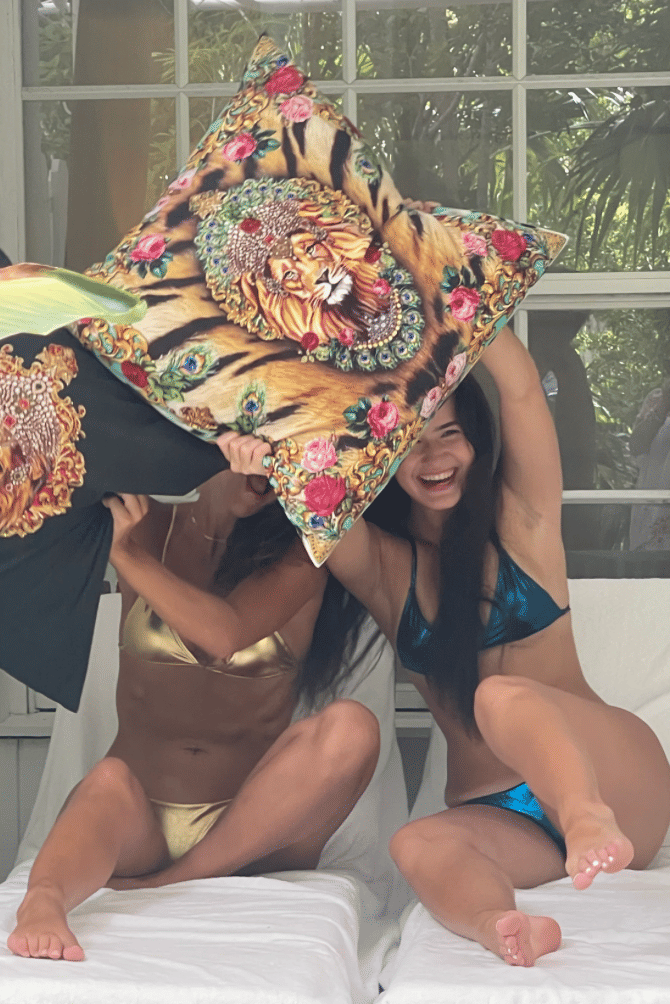 Two women in metallic bikinis are seated outdoors on white lounge chairs, playfully engaging in a pillow fight with large, decorative pillows featuring a tiger and lion print, enjoying their time at Casa Morada.