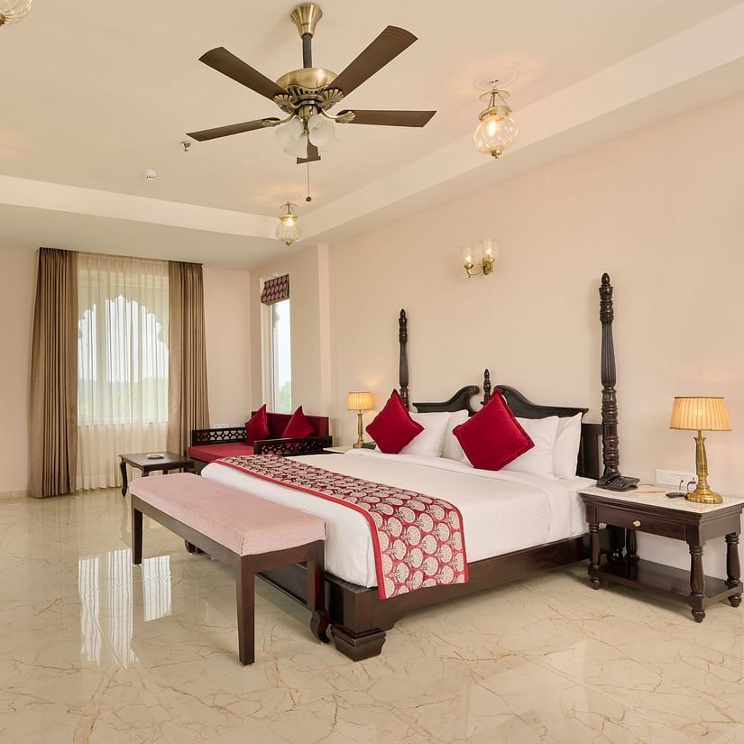 Luxurious hotel room with a four-poster bed, red accents, a seating area, and a flat-screen TV, bathed in natural light.