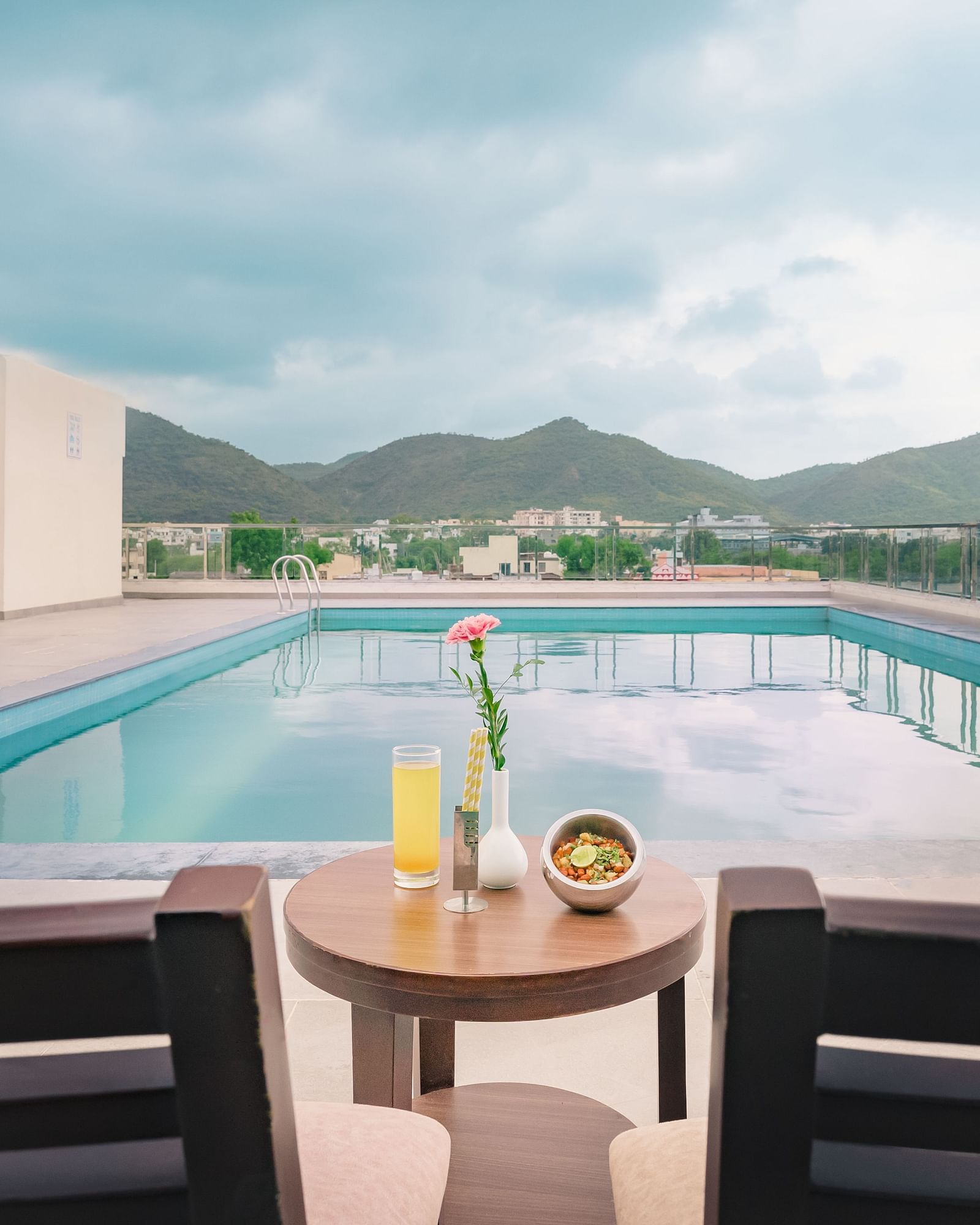 A rooftop pool at Lords Inn Udaipur offers a scenic view of distant hills, with a table set for two in the foreground.