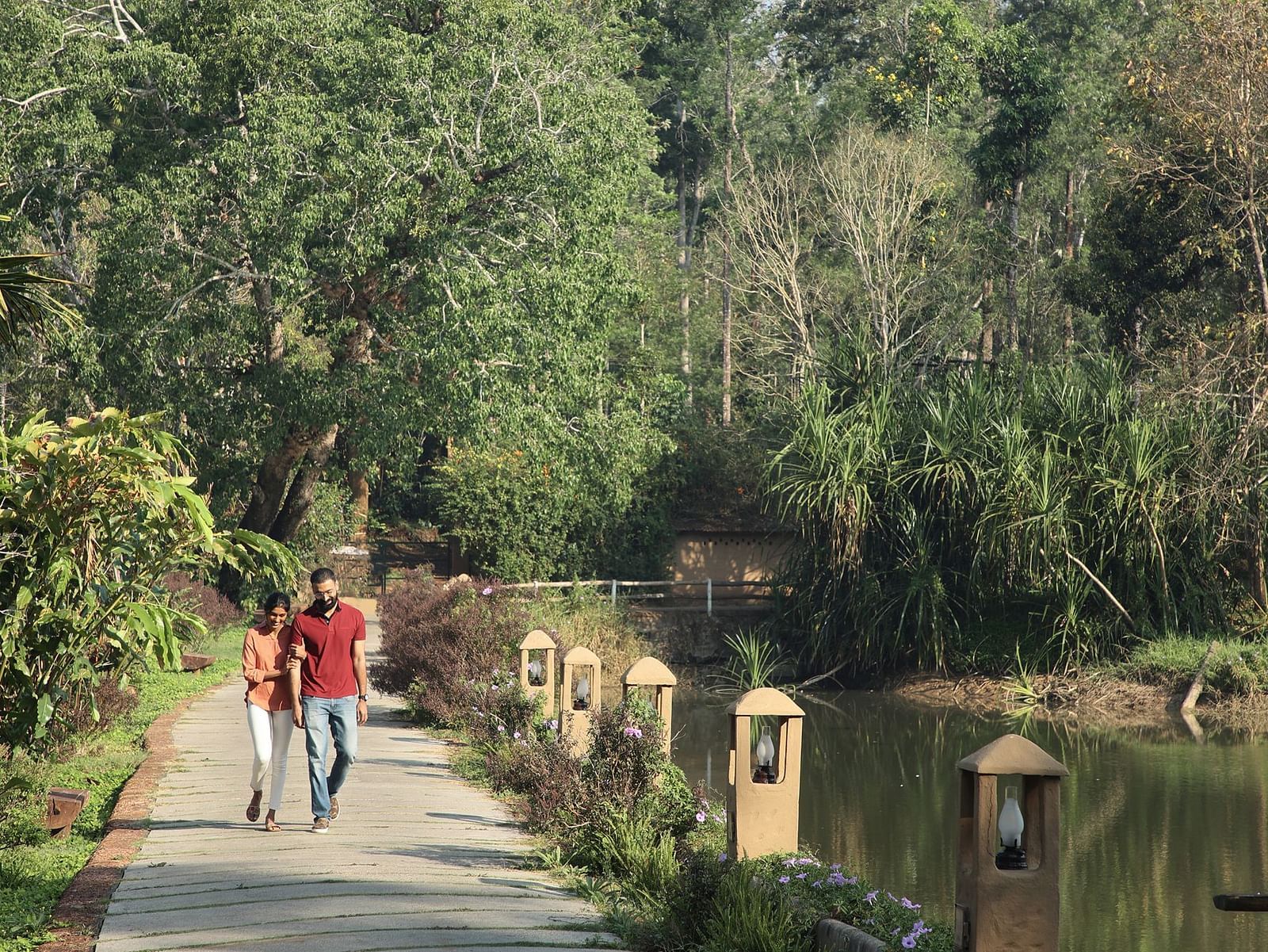 Two people walking on a path beside a pond in a lush, green park in Coorg