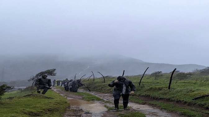 A group of people dressed in rain gear walk along a muddy path lined with green grass and wind-blown trees on a foggy, overcast day in the hills. The scene captures the wild, windswept beauty of nature.