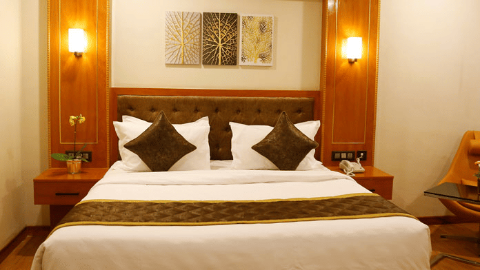 Front view of the plush double bed decorated with cushions and bed side tables on either sides of it in the Deluxe Room at Leroy Grand, Yeshwanthpur.