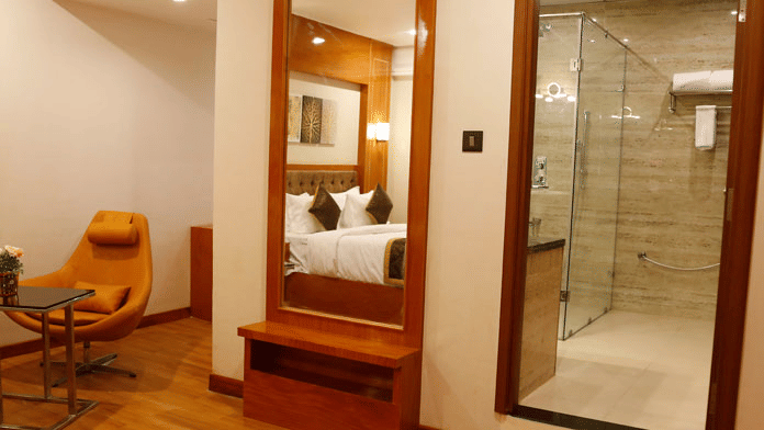 A dressing table with a full-length mirror besides the bathroom door, and a cushioned reclining chair with a coffee table placed at the Deluxe Room of Leroy Grand, Yeshwanthpur.