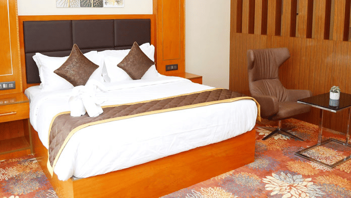 Side angle view of the double bed with a towel figurine placed over the brown bed runner at the Suite, featuring a comfortable armchair in the background at Leroy Grand, Yeshwanthpur.
