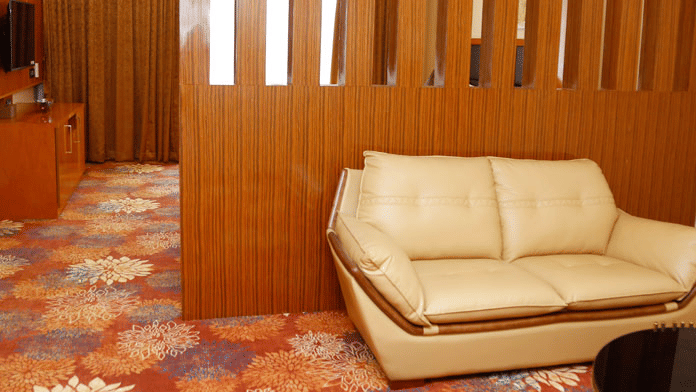 A beige-coloured leather sofa in the separate living area placed against a wooden partition in the Suite at Leroy Grand, Yeshwanthpur.