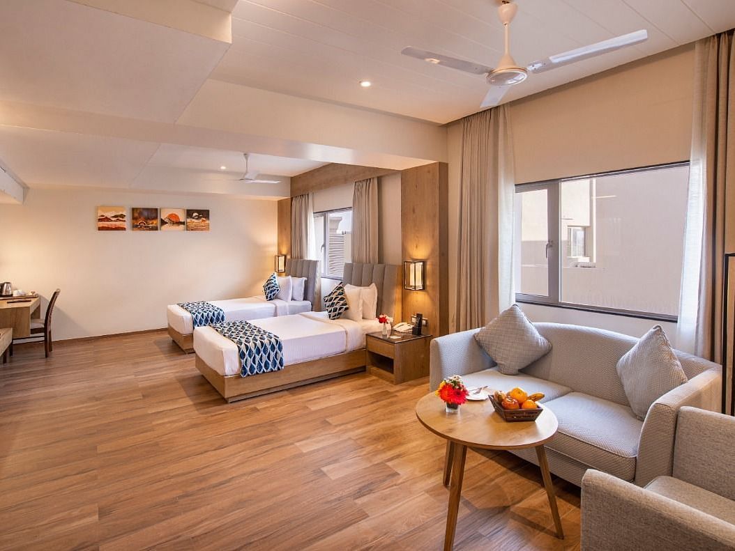 A spacious hotel suite featuring two single beds with blue and white accents, a comfortable seating area with a sofa and armchairs around a small table with fruit, a work desk with a chair, and a television, all on a wooden floor at Lords Inn Motikhavdi