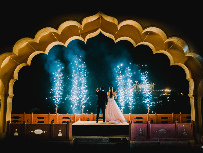 A couple stands on a raised platform under an arched structure with fireworks behind them at Tijara Fort-Palace - 19th Century, Alwar.