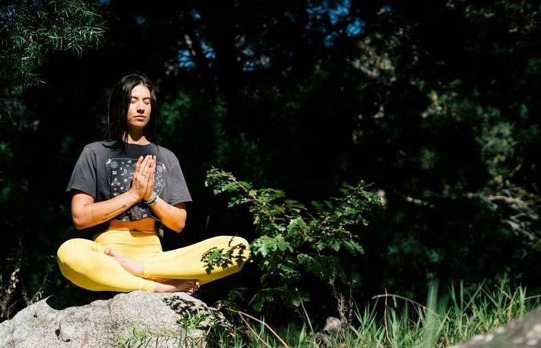 A woman with dark hair meditates peacefully in a lotus position on a rock, wearing pants and a t-shirt, surrounded by nature.