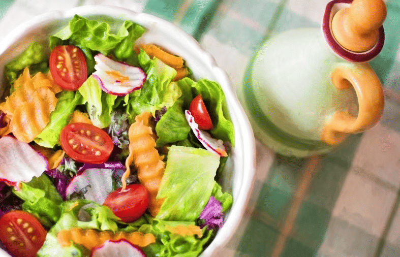 A vibrant, colorful fresh salad in a white bowl sits next to a green oil or dressing dispenser on a green and white checkered cloth at YO1 Longevity and Health Resorts, Catskills