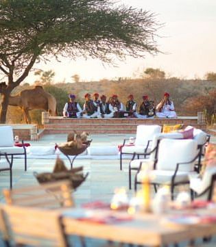 An image of outdoor seating area with seating arrangement placed in front of a podium where a group of men are sitting wearing traditional Rajasthani outfit - MANVAR Resort & Desert Camp