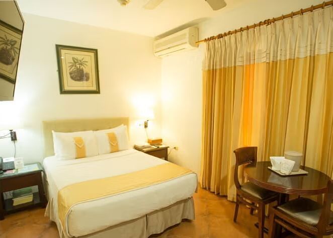 A well-lit hotel room features a bed with white linens, a nightstand, and a dining area with a table and chairs | Altamont Court, Jamaica