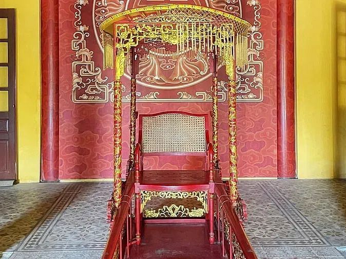 An ornate, royal red and gold throne set in an elaborately decorated interior with red pillars and traditional Chinese architectural details.