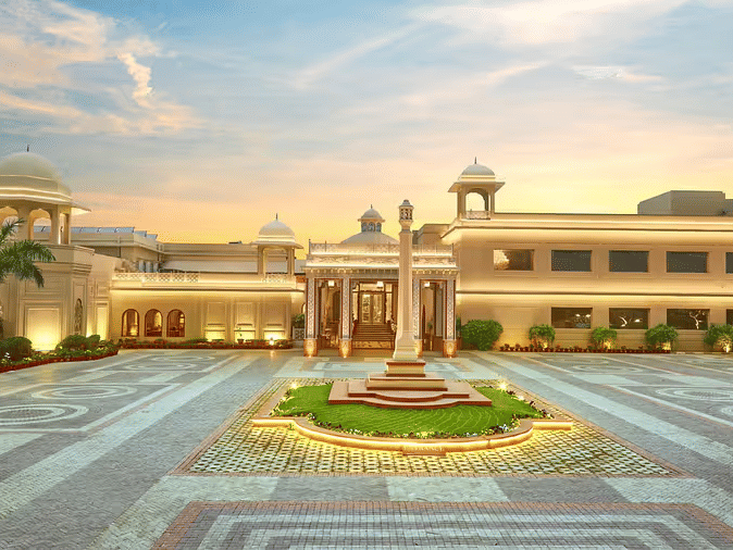 The facade view of Heritage Village Resorts & Spa, Manesar, with driveway, central fountain, and landscaped entrance under a clear sky.