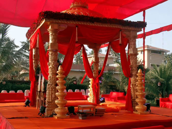 A ceremonial pavilion with decorative draping and pillars set up outdoors with palm trees in the background.