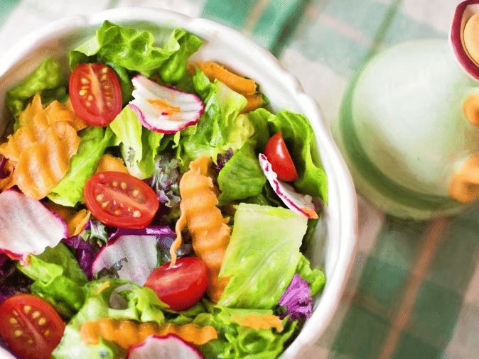 A fresh salad bowl with lettuce, cherry tomatoes, radish slices, and crinkle-cut carrots placed next to a ceramic dressing bottle.