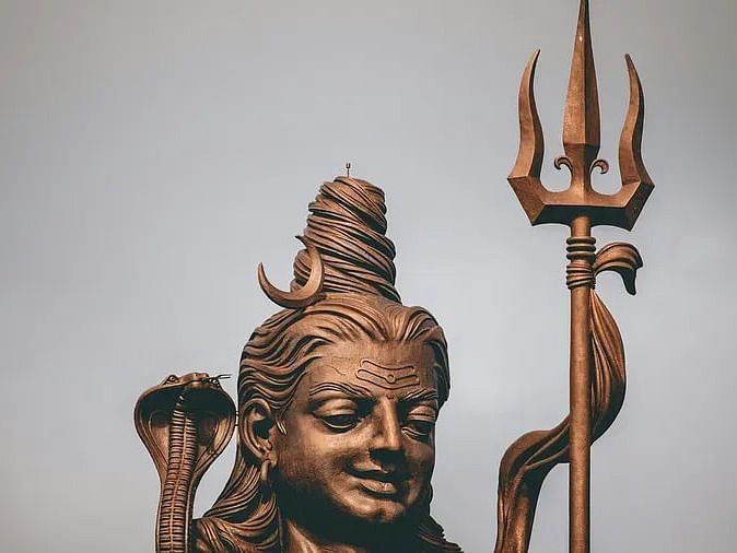 A close-up of a sculpture of Lord Shiva with detailed facial features, a flowing hairstyle, and a trident (trishul) in the background.