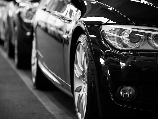 A black and white image of a few cars lined up one behind the other, with glossy reflection of the surrounding area.