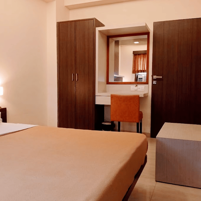 deluxe room view showcasing a beige double bed, wooden closet wardrobe, and a vanity mirror with an orange stool.