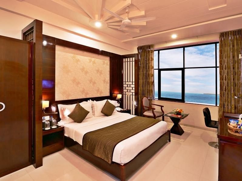 A cosy hotel room includes a king size bed with patterned bedding, a seating area near a window with a view, and warm lighting.