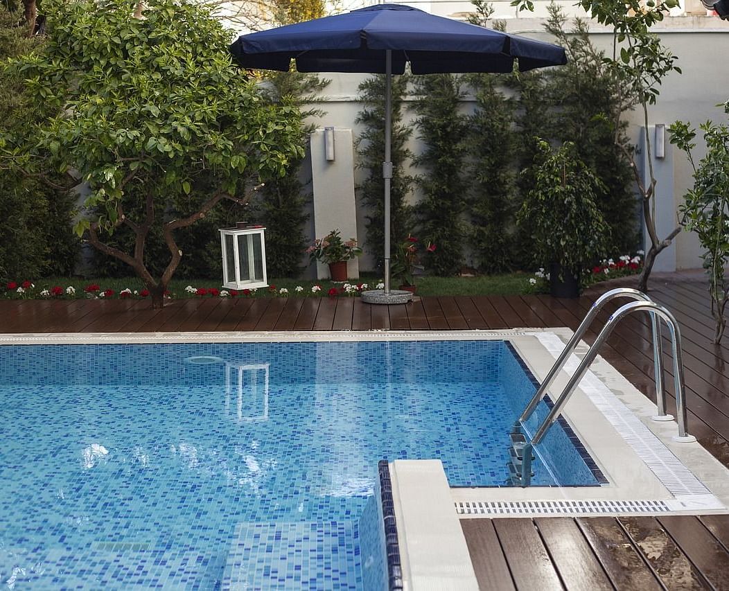 A swimming pool with a wooden deck besides it surrounded by green bushes.