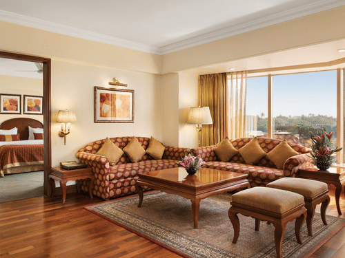 Living area of a suite with multiple sofas, a wooden centre table, lamps, and a big window with a view of the natural scenery outside.