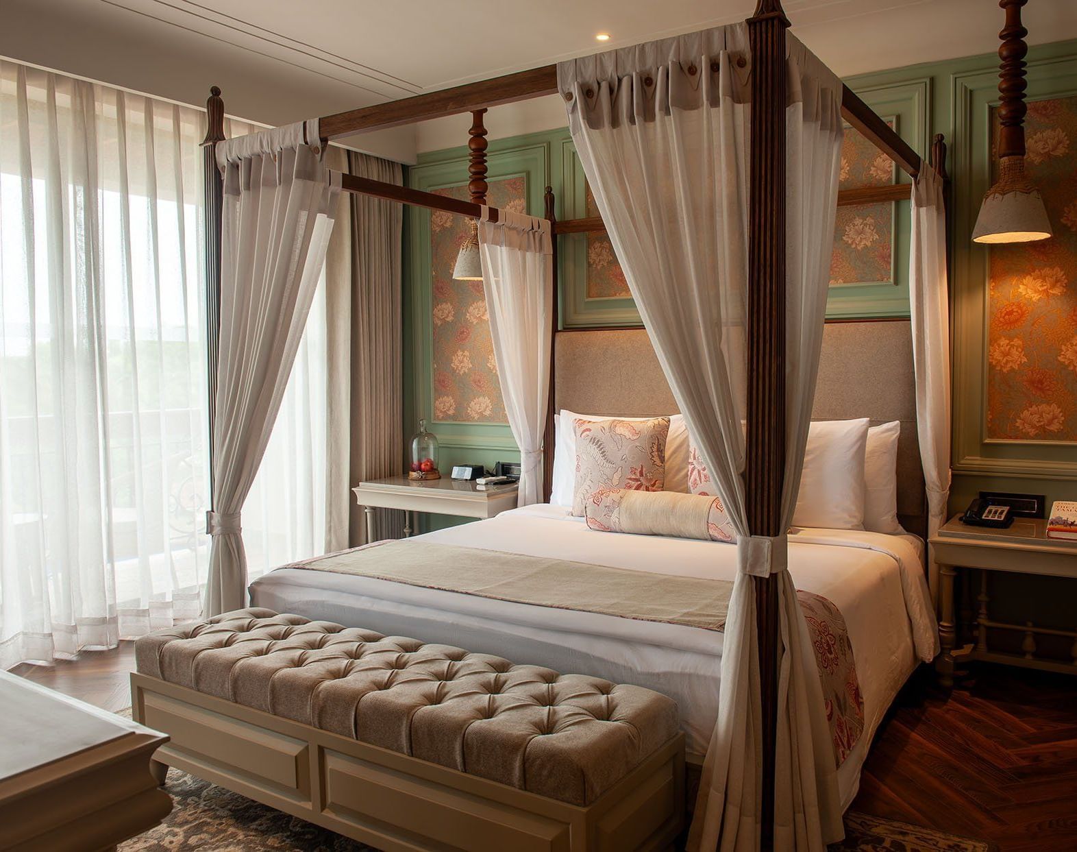 A bright and inviting suite at The Evren, Vagator, with a four-poster bed, white flowing curtains, and a plush tufted bench. The room features intricate wallpaper details and a stylish wooden floor.