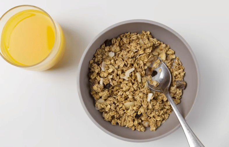 a glass of orange juice and a bowl of granola with a spoon, on a white surface.
