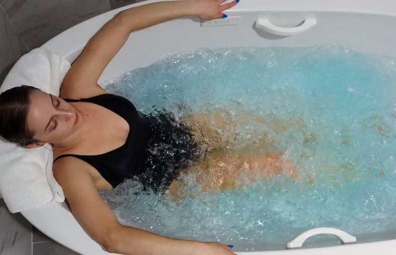 Woman in a black swimsuit enjoying a bubbling hydrotherapy tub, leaning back with eyes closed, against a marble tile background.