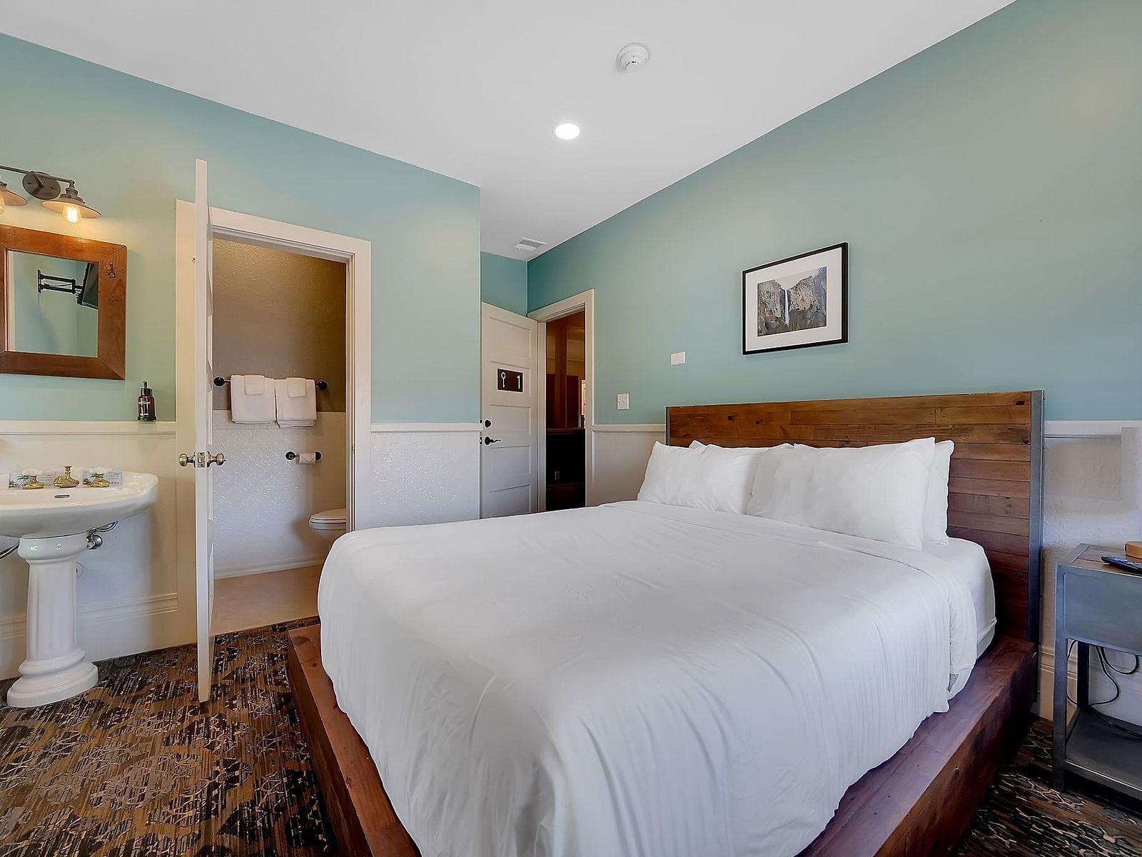 The Groveland Hotel's compact room features sage green walls, a wooden bed with crisp white bedding, and a pedestal sink with mirror.