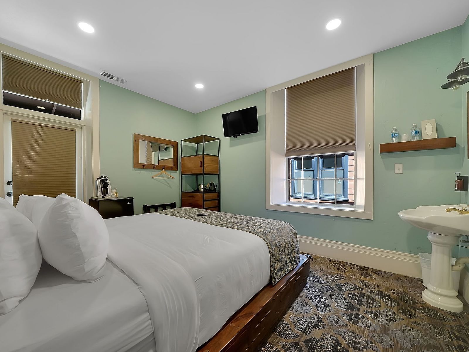 A charming bedroom at The Groveland Hotel with sage green walls, wooden platform bed, pedestal sink, and patterned area rug throughout.