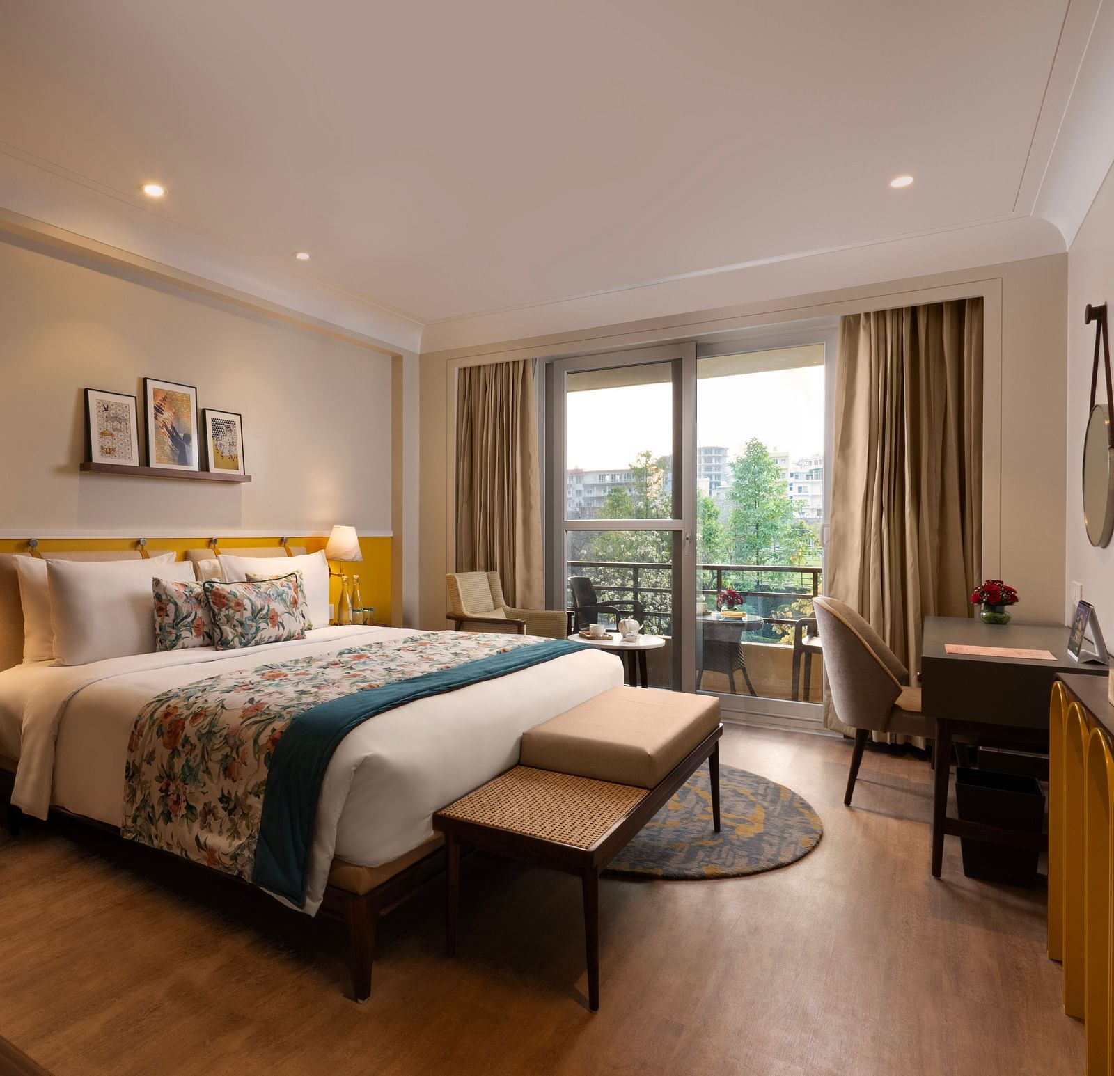 A spacious interior of room with a large bed, a sitting area with a sofa and a coffee table, and a desk. The room has large windows with curtains and a balcony overlooking a view.