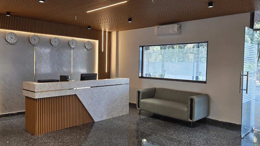 A cosy reception lounge with a wooden counter, grey sofa, and large glass windows letting in natural light.