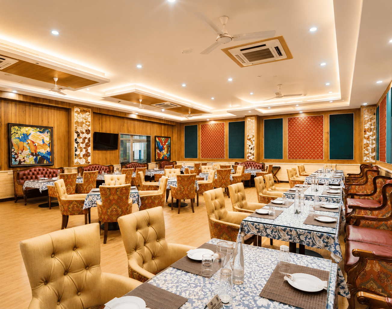 Teapot restaurant with tufted cream chairs, red leather sofas, printed tablecloths and parrot artwork on panelled walls at MAYFAIR World Cup Village, Rourkela.