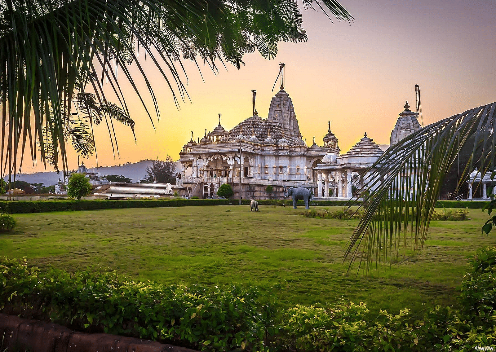 An overview of a Jain temple with the sun setting behind it and a garden area in the foreground.